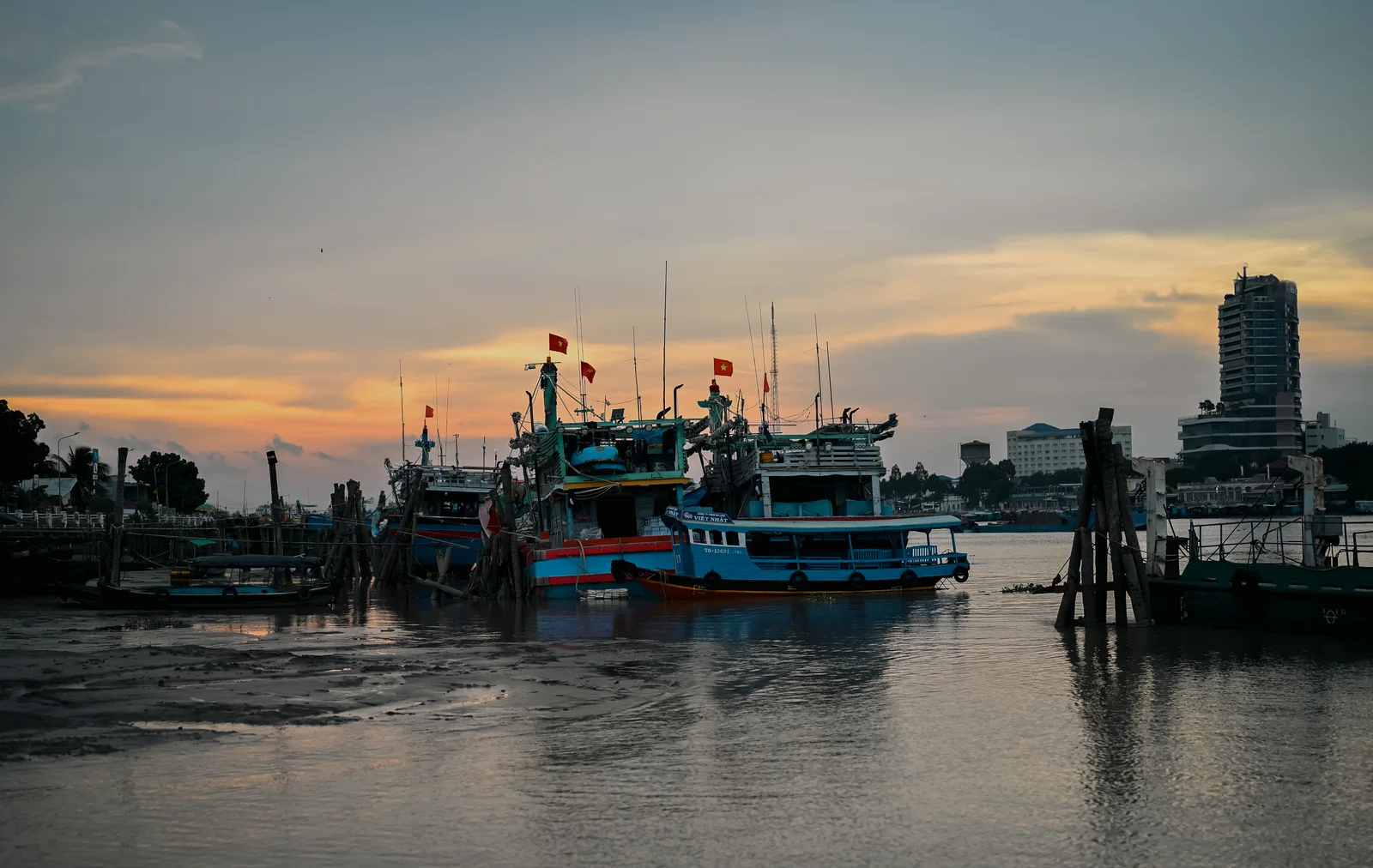 Fishing boats and river ferries moored along the riverbank at twilight with city skyline