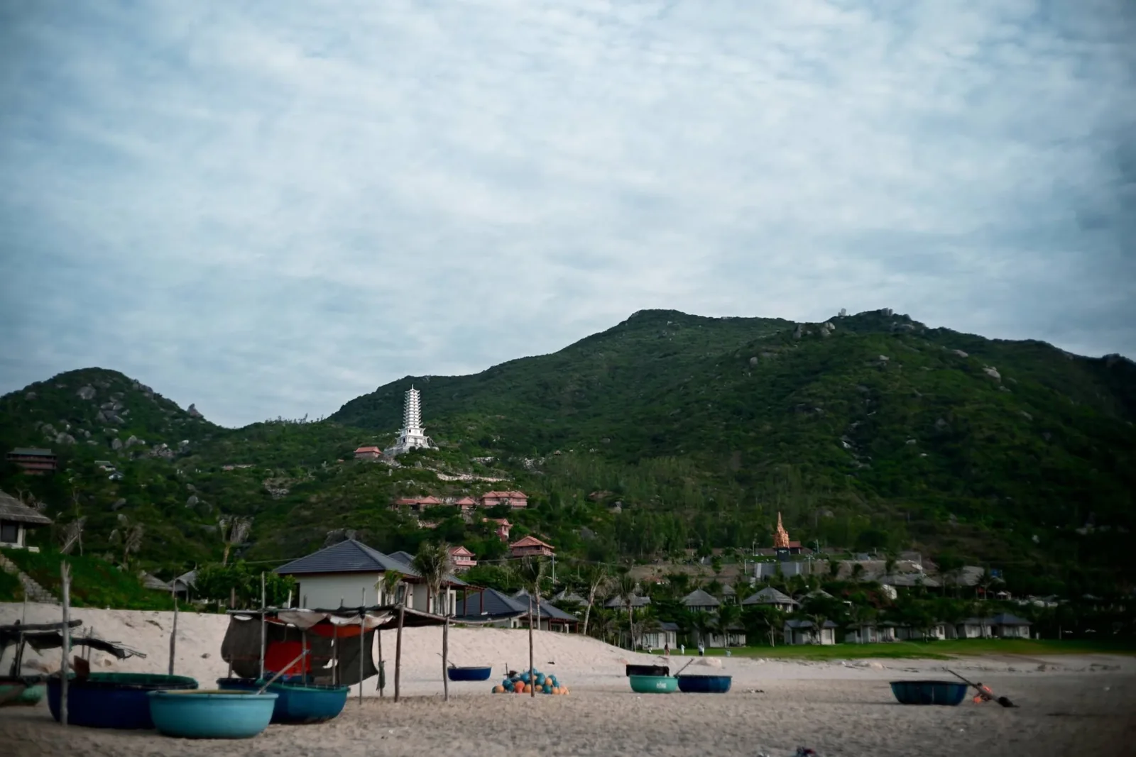 Traditional coracle boats resting on golden sand overlooking Quy Nhơn bay with Linh Phong seated Buddha atop the distant mountain
