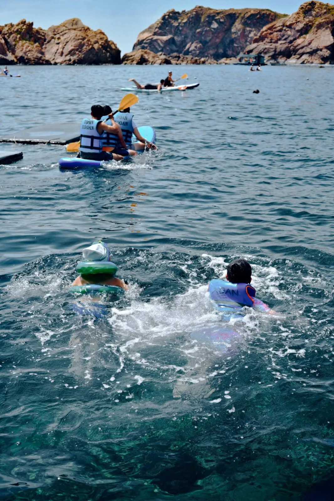 Swimmers in life jackets and SUP paddlers floating in turquoise waters beside towering coastal sea cliffs in Quy Nhơn