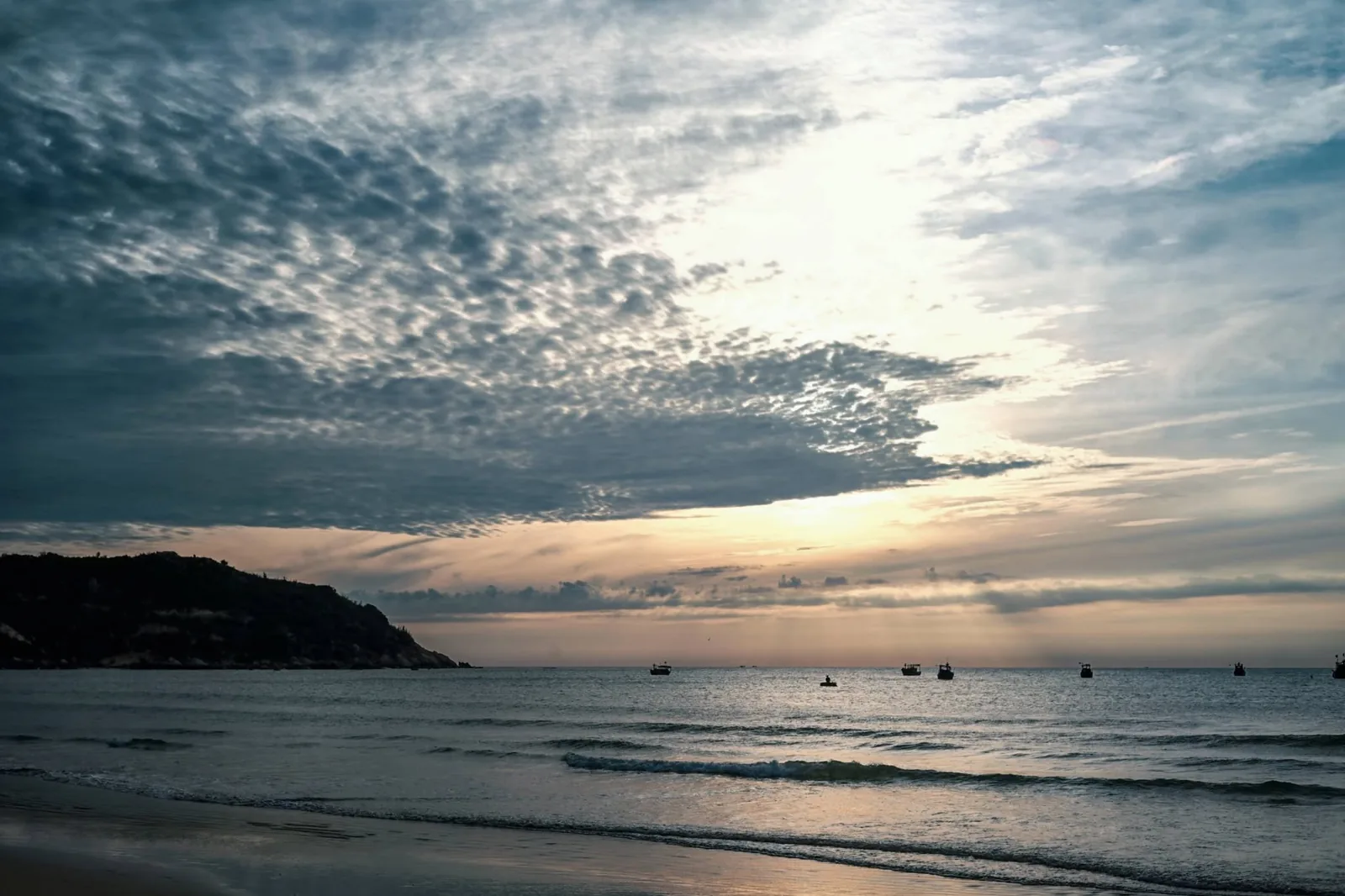 Sunrise over Quy Nhơn sea with golden rays breaking through dramatic clouds above distant fishing boats