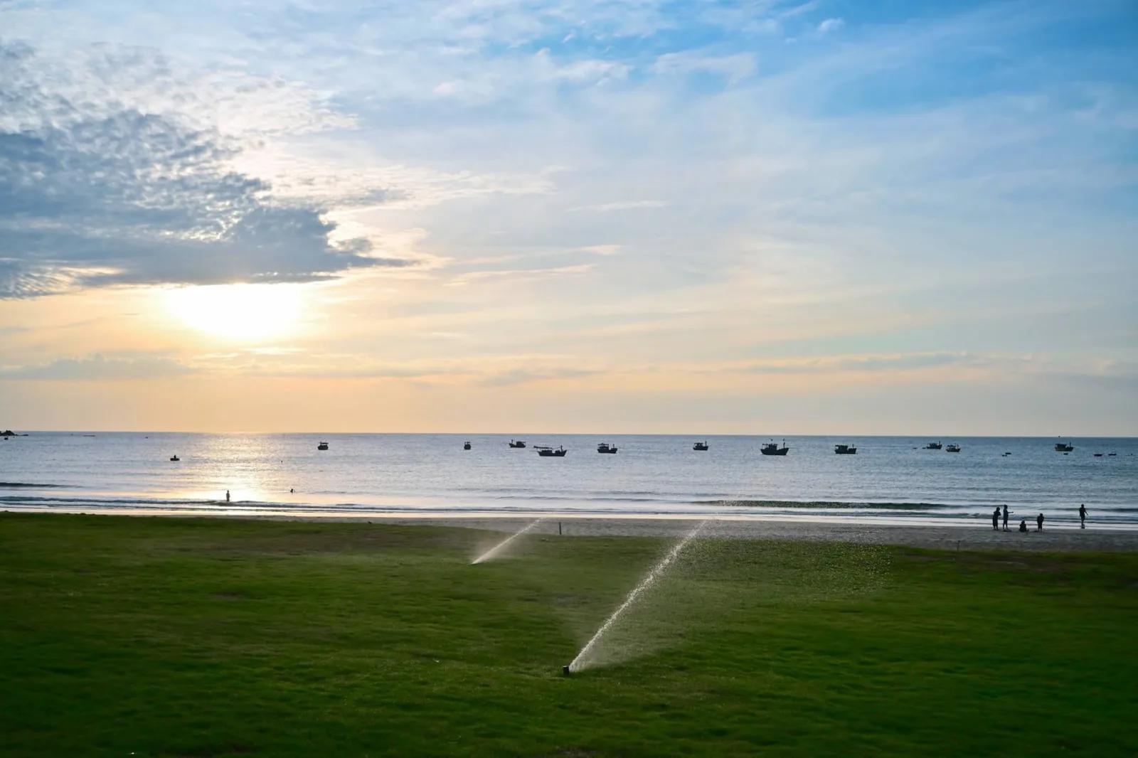 Lawn sprinklers casting water droplets in the golden morning light over Quy Nhơn beach and offshore boats