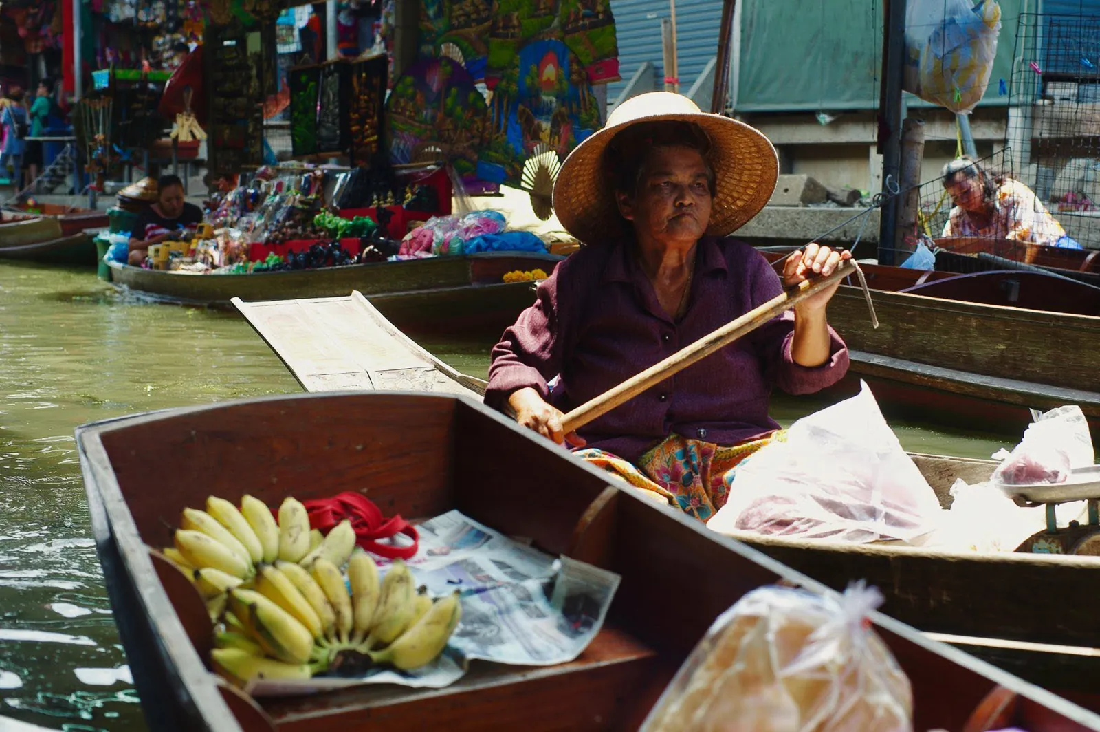 Vendor wearing woven conical hat rowing a wooden boat filled with ripe bananas