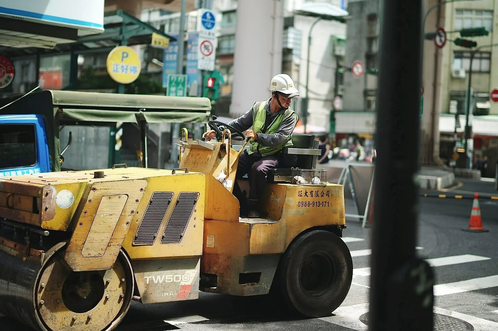 Road construction worker in safety vest and hardhat operating heavy paving machinery at a street corner